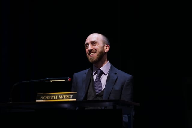 Adam wears a shirt and tie, with a confident smile in the National Theatre's My Country