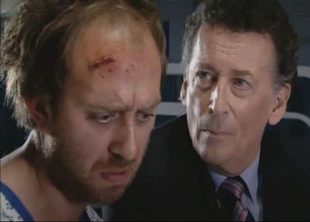 Adam looks despondent, with a cut across his head while a man looks on him