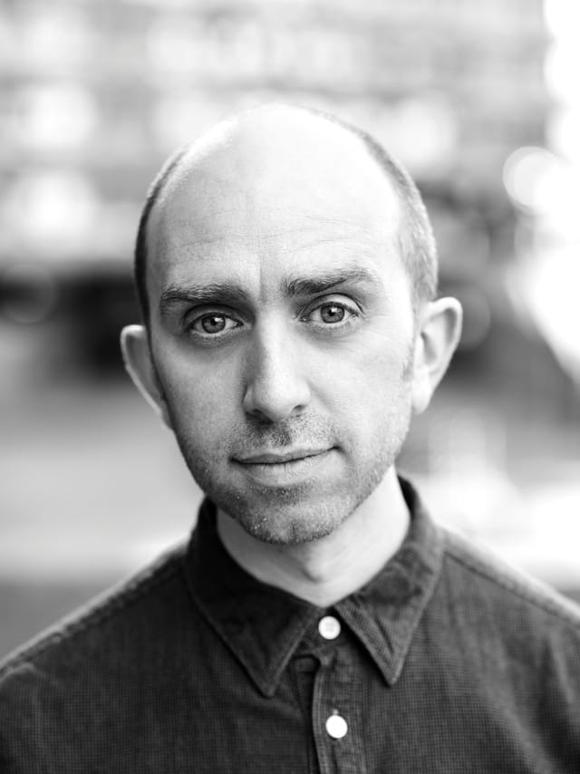 A black and white portrait shot of Adam, wearing a dark shirt and gently smiling