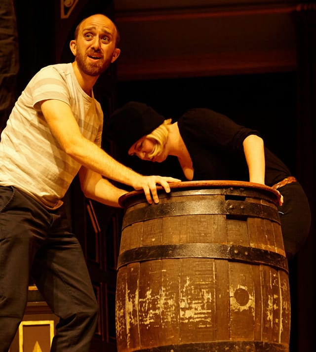 Adam looks out into the audience wearing a light t-shirt, whilst leaning over a barrel