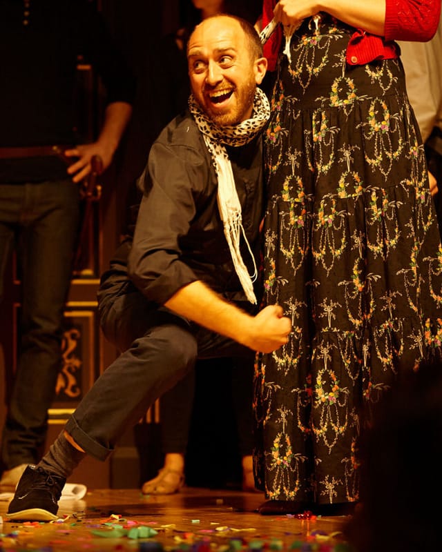 Adam crouches down around someone wearing a dark patterned skirt. He is smiling.