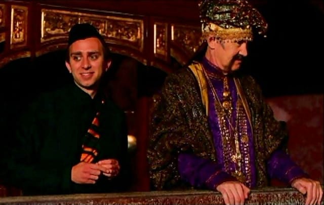 Adam stands behind a man in a bright, magicians outfit, looking intrigued