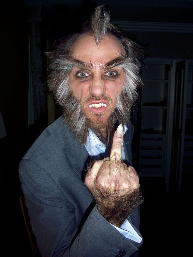Adam wears prosthetic teeth, and a short spikey grey wig whilst gesturing with his middle finger
