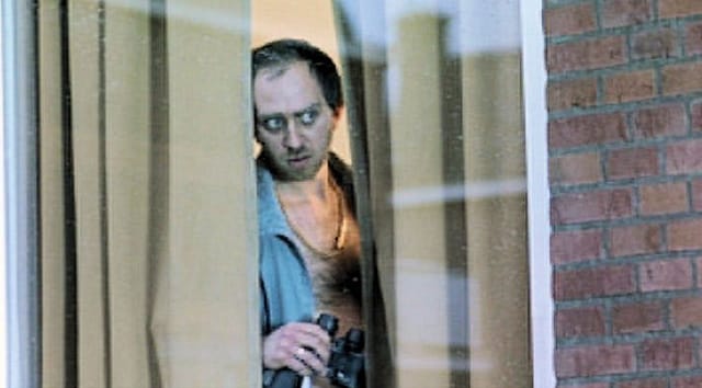 Adam holds a pair of binoculars while staring out a window from behind a curtain